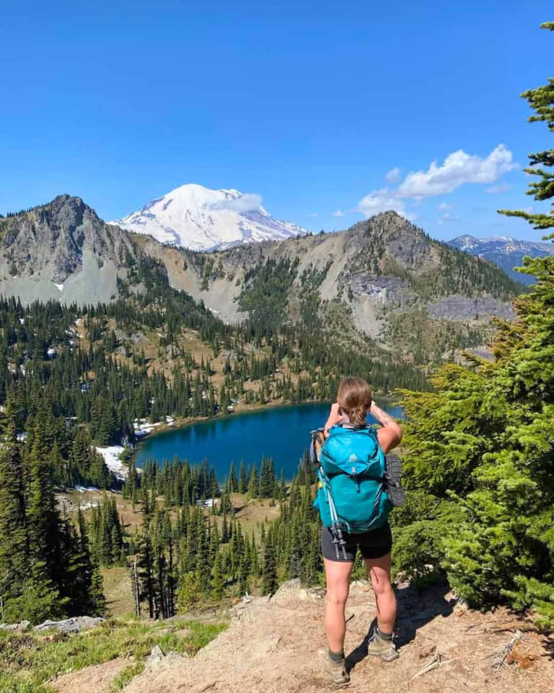 A sturdy, lightweight, and comfortable backpacking backpack is one of the most important parts of gear. A woman hiking who is taking a photo of Mt. Rainier on a beautiful summer, sunny day.