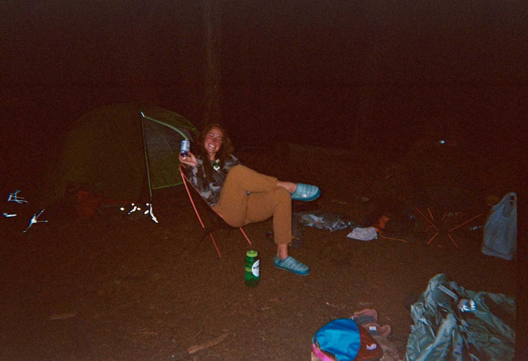 A polaroid photo of a woman wearing outdoor attire and sitting in a foldable chair at their backpacking camp for the night.