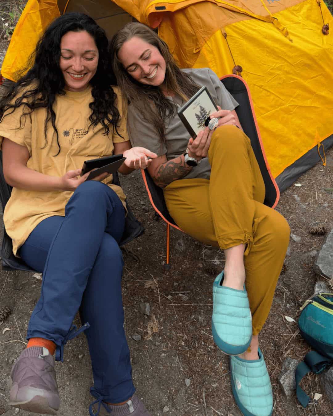 Two women reading at camp after a long day of backpacking in their foldable chairs with their Coalatree Trailhead pants on.
