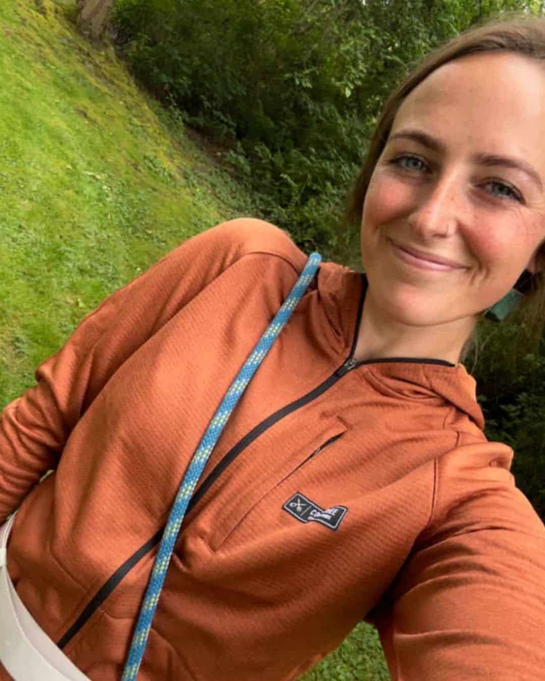 A woman wearing a burnt orange color Coalatree Baseline Midlayer jacket while walking her dog on a grassy trail.