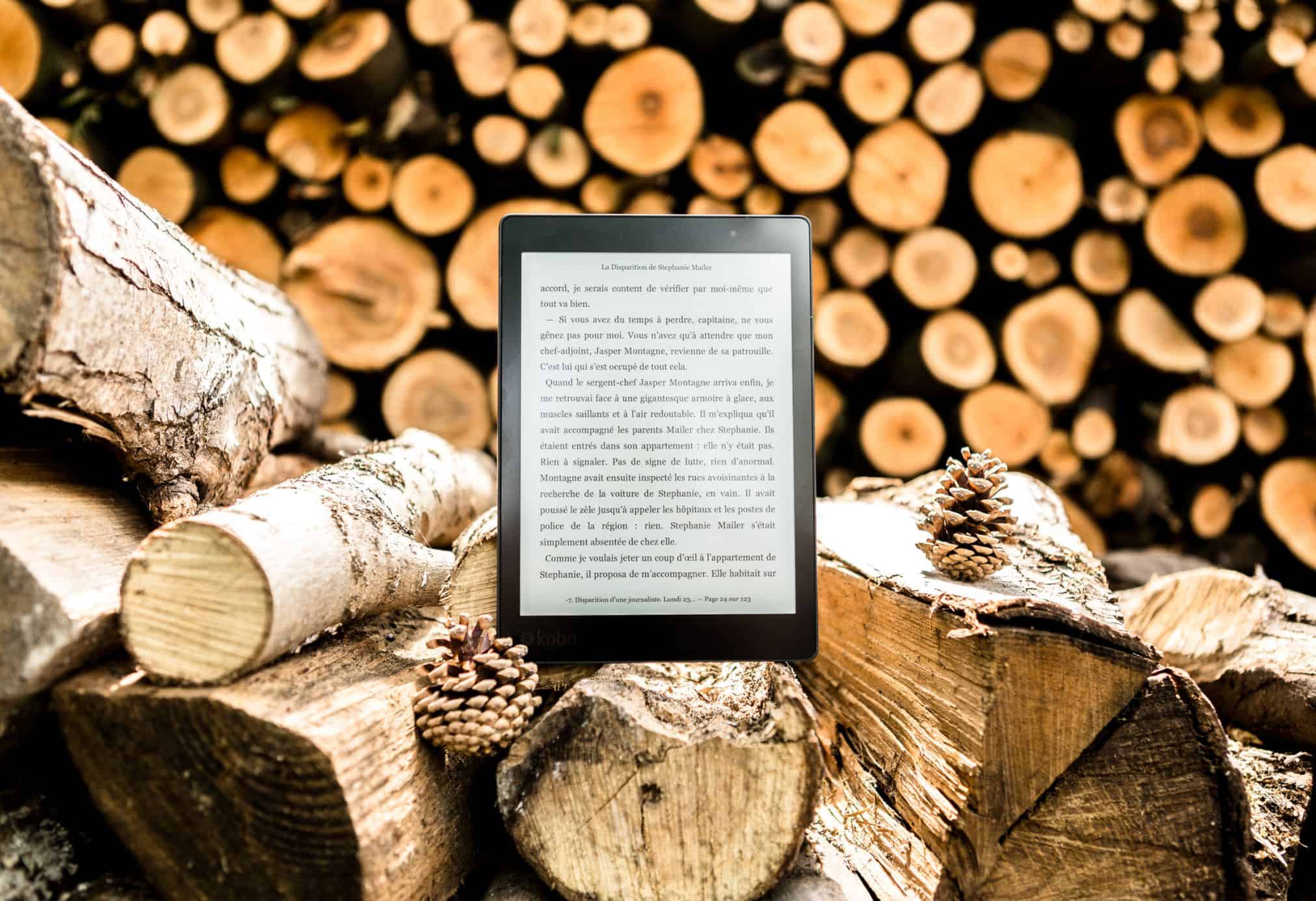 A Kindle sitting on a beautiful pile of logs with logs in the background showcasing how easy a Kindle is to use.