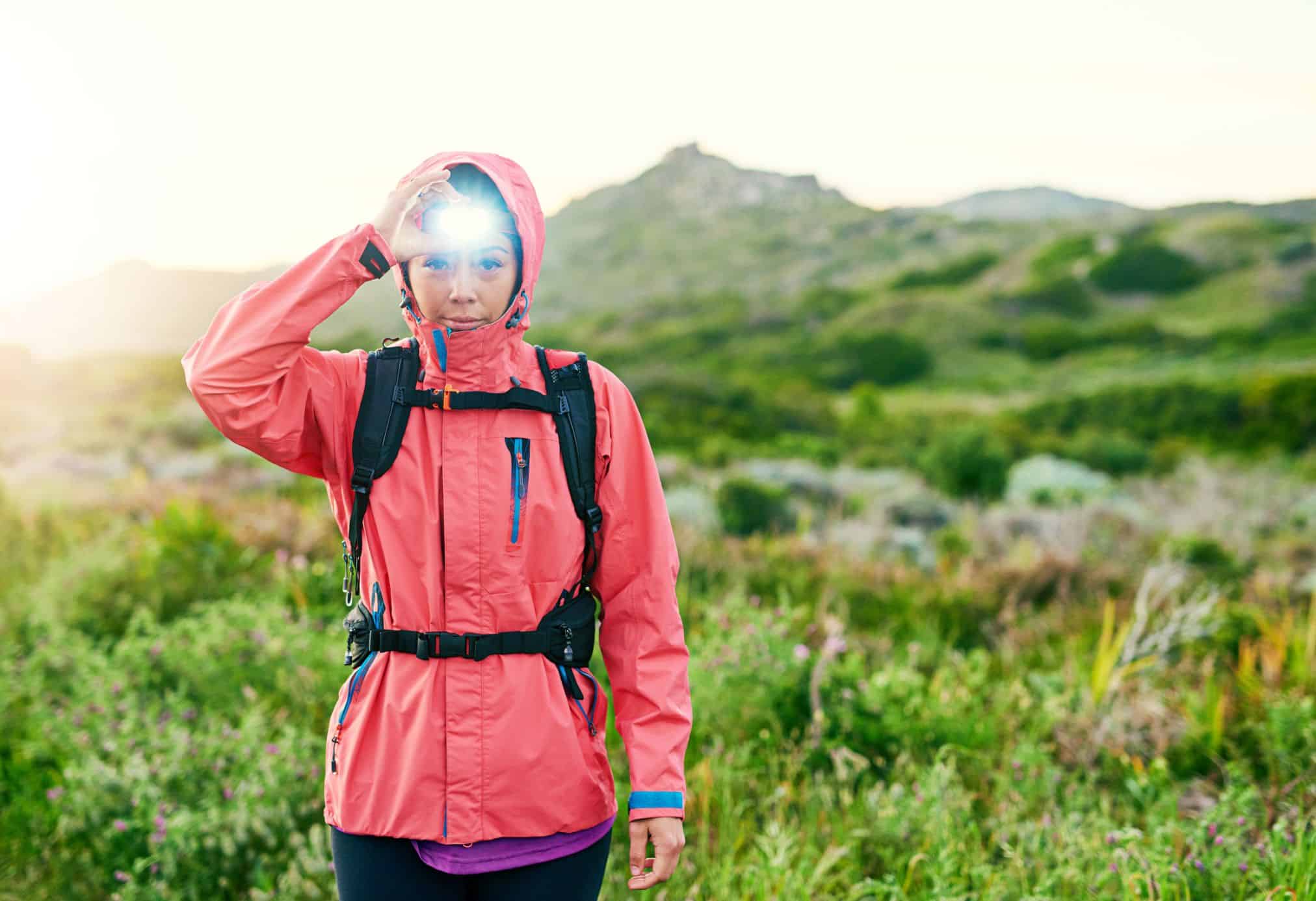 A woman in a pink jacket with a headlight on and hiking. Headlamps are one of the best gifts for road trips, you never know when you may need one.