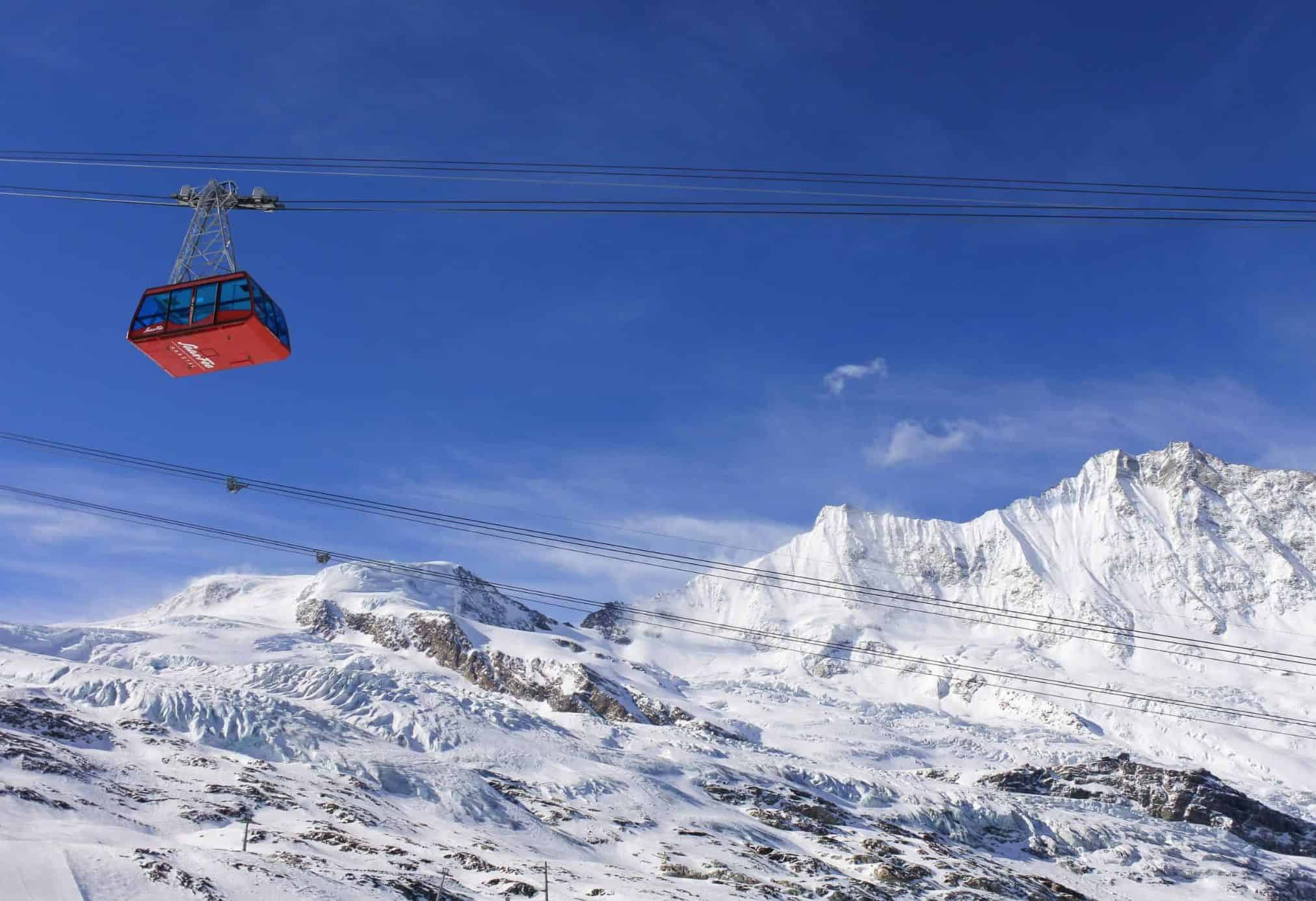 A red gondola going into the mountains in Saas-Fee with snow on the mountains in the background and a blue sky.