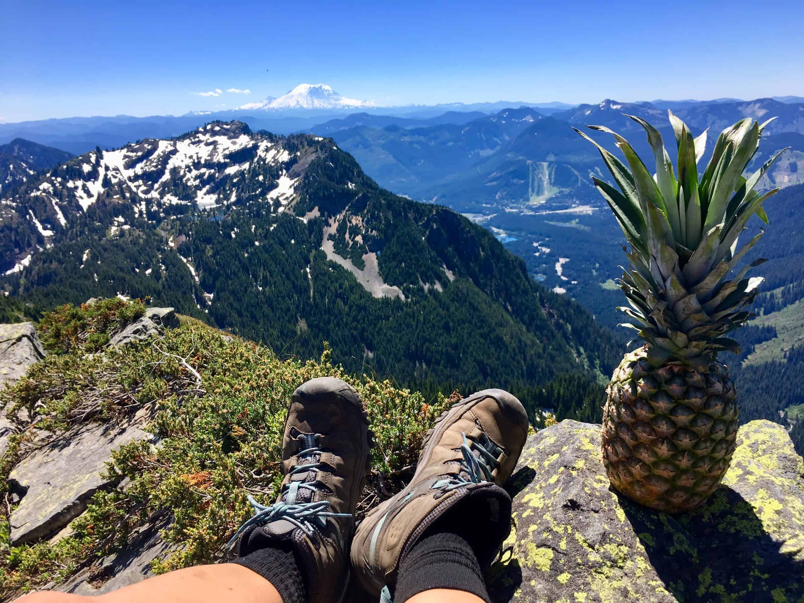 A fun photo of a pair of hiking boots on top of a mountain with Mount Rainier in the background and a pineapple.