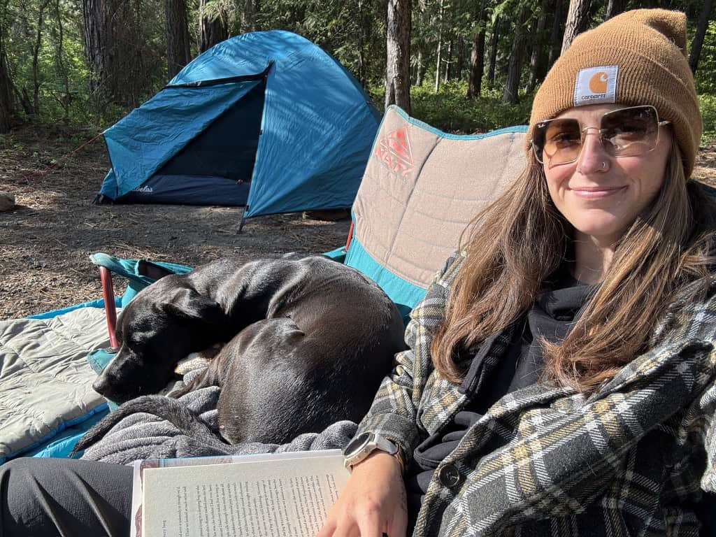 A woman reading at a campground with er dog while sitting in a beanie and flannel with hiking pants.